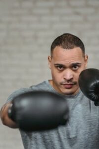 Confident young strong ethnic male boxer in activewear and gloves punching and looking at camera during workout in boxing club