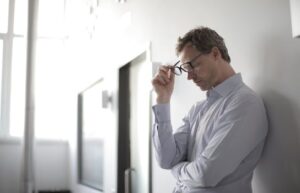 Thoughtful man in a bright room holding his glasses while leaning against a wall. What Is Nightfall and Why It Happens Frequently: Causes, Effects, and Solutions – Meditation helps control nightfall naturally