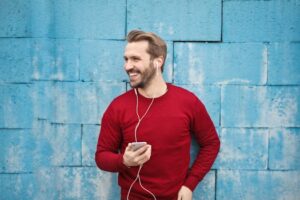 A joyful man in a red sweater smiles while listening to music on his smartphone against a blue brick wall. What Is Nightfall and Why It Happens Frequently: Causes, Effects, and Solutions – Meditation helps control nightfall naturally